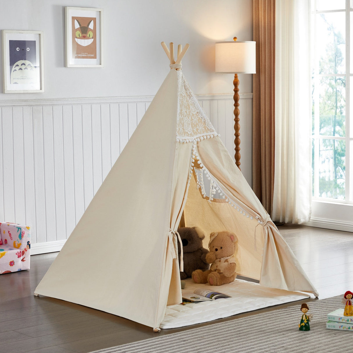 Beige children's teepee with teddy bear and books in a room with a lamp and window.